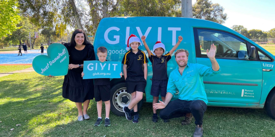 A male GIVIT staff member crouching next to three young schoolboys wearing santa hats and holding GIVIT signs, plus their female teacher.