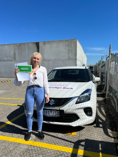 A young woman smiling standing in front fo a car holding her learner license.